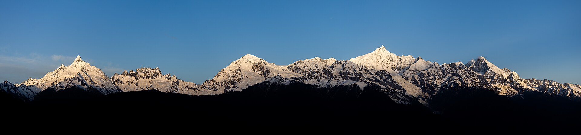 梅里雪山群峰全景:白雪覆盖的山脊在湛蓝天空下连绵起伏,山脚隐入深色剪影